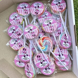 May include: A cardboard box containing numerous wrapped lollipops. Each lollipop features a pink and white striped design, a Minnie Mouse graphic, and the text "Happy 1st Birthday Neyrer". One lollipop has a swirl design.