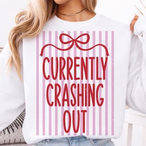 May include: White sweatshirt with the text "CURRENTLY CRASHING OUT" in red, set against a pink and white striped background. The design includes a red bow at the top, creating a gift box appearance.