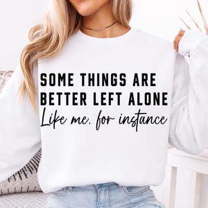 Puede incluir: Sudadera blanca con el texto negro "SOME THINGS ARE BETTER LEFT ALONE Like me. for instance". La sudadera tiene mangas largas y cuello redondo. La modelo lleva vaqueros azul claro.