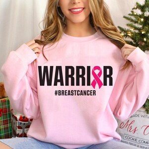 May include: A light pink sweatshirt with the word "WARRIOR" in bold black letters and a pink ribbon graphic. Below the word is the hashtag "#BREASTCANCER". The sweatshirt is a crew neck style.