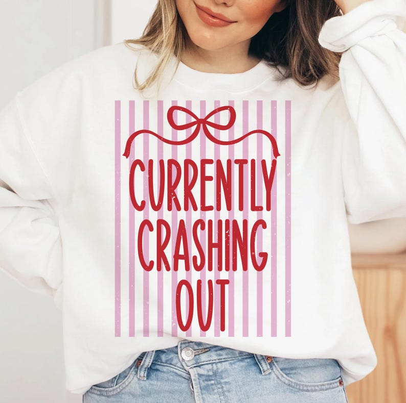 May include: White sweatshirt with the text "CURRENTLY CRASHING OUT" in red, set against a pink and white striped background. The design includes a red bow at the top. The sweatshirt is a casual, comfortable style.