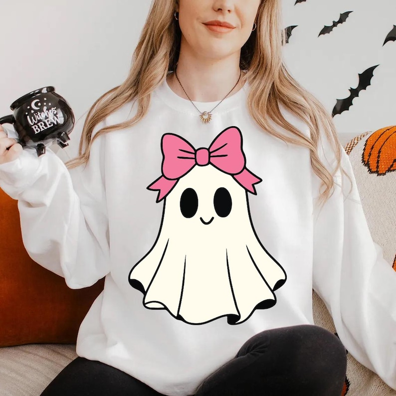 K&ouml;nnte beinhalten: Wei&szlig;es Sweatshirt mit einem Cartoon-Geist-Design und einer rosa Schleife. Der Geist hat schwarze Augen und einen l&auml;chelnden Mund. Das Sweatshirt ist Teil einer Halloween-Kollektion.