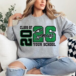 Pode incluir: Uma camisola cinzenta com o texto "CLASS OF 2026 Senior YOUR SCHOOL" em preto e verde. A camisola &eacute; usada por uma pessoa sentada num sof&aacute; branco, usando cal&ccedil;as de ganga azuis.