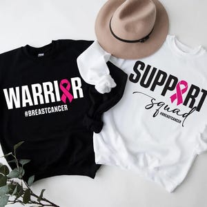 May include: Two sweatshirts, one black and one white, with breast cancer awareness themes. The black sweatshirt says "WARRIOR" with a pink ribbon and "#BREASTCANCER". The white sweatshirt says "SUPPORT squad" with a pink ribbon and "#BREASTCANCER". A tan hat rests on top.