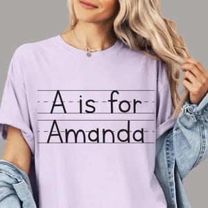 May include: A light purple t-shirt with the text "A is for Amanda" in black. The text is written in a school-style font, with lines above and below the text. The shirt is a short-sleeved crew neck.