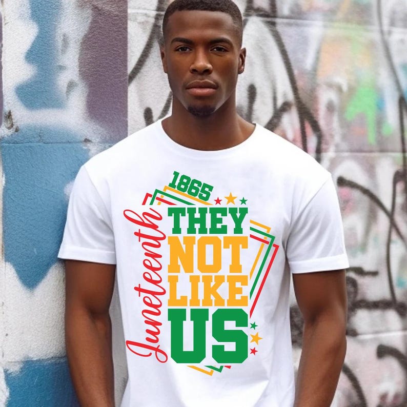 May include: White t-shirt with the text "Juneteenth 1865 THEY NOT LIKE US" in red, green, and yellow. The design includes stars and geometric shapes. The shirt is worn by a person in front of a graffiti wall.