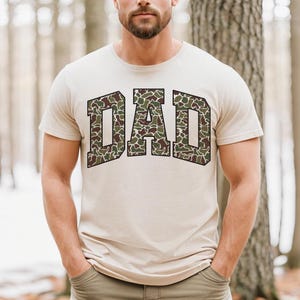 May include: A beige t-shirt with a camouflage pattern spelling out "DAD" in a bold, blocky font.