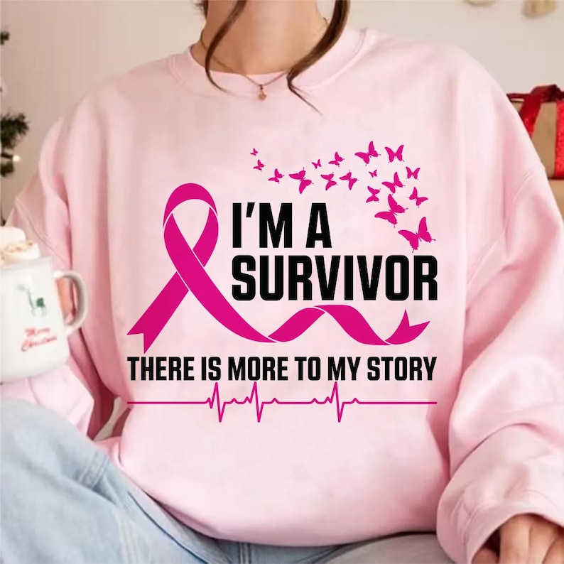 以下が含まれることがあります： ピンク色のスウェットシャツ。ピンクのリボンと黒字で「I'M A SURVIVOR」と書かれています。デザインにはピンクの蝶と「THERE IS MORE TO MY STORY」の文字、心拍ラインが含まれています。
