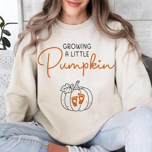 Op de afbeelding: Crèmekleurige sweatshirt met de tekst "GROWING A LITTLE Pumpkin" in oranje script. Een zwart-witte pompoenafbeelding met oranje babyvoetjes staat eronder. De sweatshirt heeft een casual stijl.