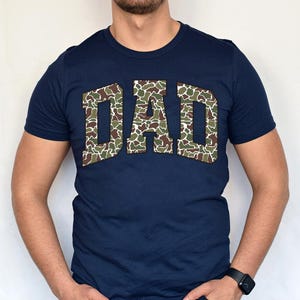 May include: A navy blue t-shirt with a large "DAD" graphic in a camouflage pattern.