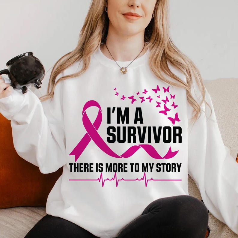 以下が含まれることがあります： ピンクのリボン、蝶、そして黒で「I'M A SURVIVOR THERE IS MORE TO MY STORY」というテキストが入った白いスウェットシャツ。心拍数の線グラフが下にあります。スウェットシャツはカジュアルなスタイルです。