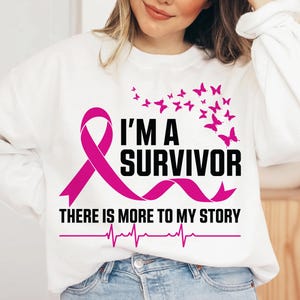 以下が含まれることがあります： ピンクのリボンと「I'M A SURVIVOR」の黒い文字が入った白いスウェットシャツ。ピンクの蝶がテキストの上にあります。テキストの下には「THERE IS MORE TO MY STORY」と心拍ラインがあります。