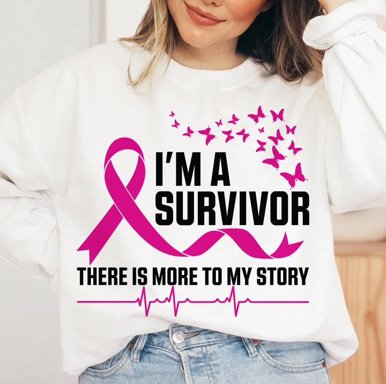 以下が含まれることがあります： ピンクのリボンと「I'M A SURVIVOR」の黒い文字が入った白いスウェットシャツ。ピンクの蝶がテキストの上にあります。テキストの下には「THERE IS MORE TO MY STORY」と心拍ラインがあります。
