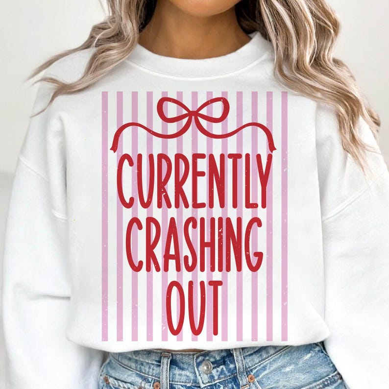 May include: White sweatshirt with a red bow and the text "CURRENTLY CRASHING OUT" in red. The text is set against a background of vertical pink and white stripes. The sweatshirt is a casual style.