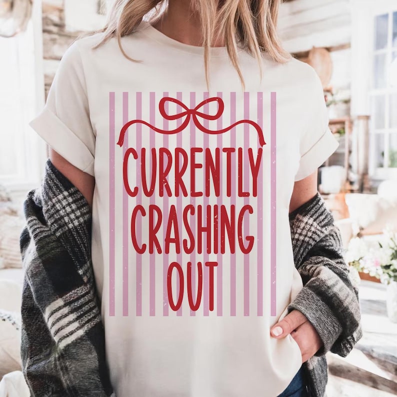 May include: Cream-colored t-shirt with the text "CURRENTLY CRASHING OUT" in red, framed by a red bow and vertical pink stripes. The shirt has short sleeves and a relaxed fit.