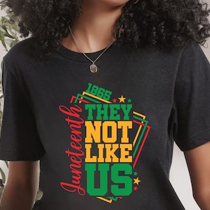 May include: Black t-shirt with a colorful graphic design. The design includes the words "Juneteenth", "1865", "THEY", "NOT", "LIKE", and "US" in green, yellow, and red. The shirt is a crew neck.