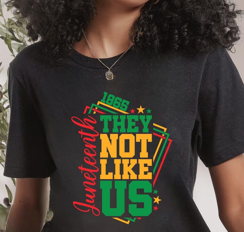 May include: Black t-shirt with a colorful graphic design. The design includes the words "Juneteenth", "1865", "THEY", "NOT", "LIKE", and "US" in green, yellow, and red. The shirt is a crew neck.