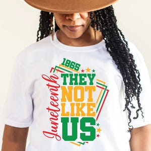 May include: White t-shirt with a colorful graphic design. The design includes the word "Juneteenth" in red, and the phrase "THEY NOT LIKE US" in green and yellow, with the year "1865" in green. The shirt is worn by a person wearing a brown hat.
