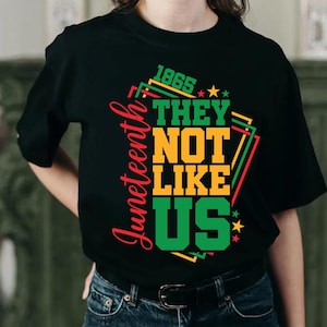 May include: Black t-shirt with the text "1865 THEY NOT LIKE US" in green, yellow, and red. The word "Juneteenth" is written vertically in red on the left side. The shirt has a crew neck and short sleeves.