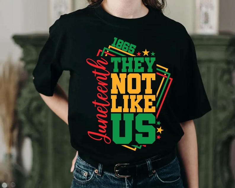 May include: Black t-shirt with the text "1865 THEY NOT LIKE US" in green, yellow, and red. The word "Juneteenth" is written vertically in red on the left side. The shirt has a crew neck and short sleeves.