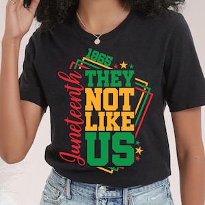 May include: Black t-shirt with a graphic design celebrating Juneteenth. The design features the text "1865 THEY NOT LIKE US" in green and yellow, with the word "Juneteenth" in red. Stars and geometric shapes complete the design.