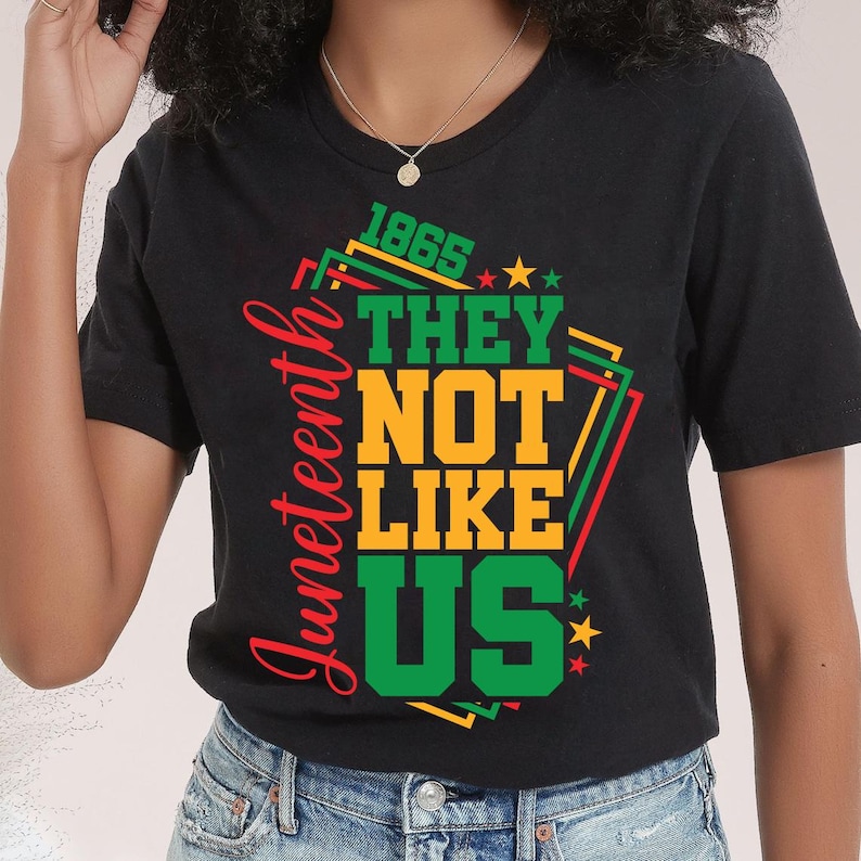 May include: Black t-shirt with a graphic design celebrating Juneteenth. The design features the text "1865 THEY NOT LIKE US" in green and yellow, with the word "Juneteenth" in red. Stars and geometric shapes complete the design.