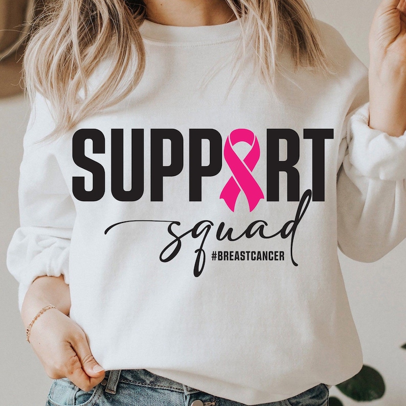 May include: White sweatshirt with the words "SUPPORT SQUAD" in black block letters and a pink ribbon graphic. Below, the word "squad" is written in a cursive font, with the hashtag "#BREASTCANCER" underneath.
