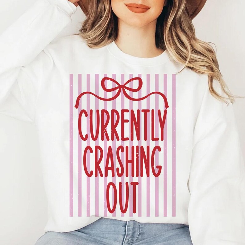 May include: White sweatshirt with a red and pink striped graphic. The graphic features a red bow and the words "CURRENTLY CRASHING OUT" in a bold, red font. The sweatshirt is a casual, comfortable style.