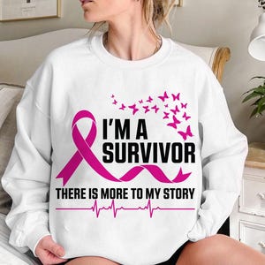 以下が含まれることがあります： ピンクのリボンと「I'M A SURVIVOR THERE IS MORE TO MY STORY」の文字が入った白いスウェットシャツ。ピンクの蝶と心拍ラインも特徴です。
