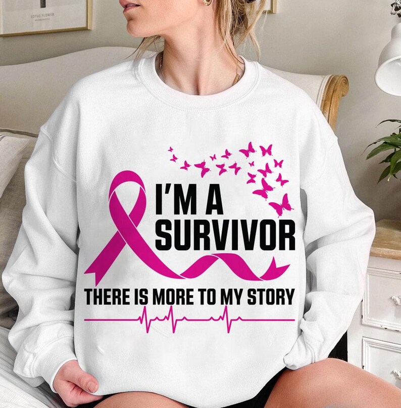 以下が含まれることがあります： ピンクのリボンと「I'M A SURVIVOR THERE IS MORE TO MY STORY」の文字が入った白いスウェットシャツ。ピンクの蝶と心拍ラインも特徴です。