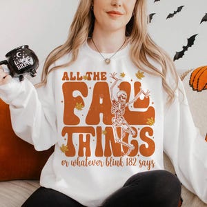 Puede incluir: Sudadera blanca con el texto "ALL THE FALL THINGS or whatever blink 182 says" en naranja. El diseño incluye un esqueleto y hojas de otoño. La sudadera es perfecta para la temporada de otoño y Halloween.