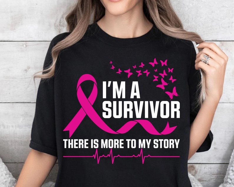以下が含まれることがあります： ピンクのリボンと白い文字で「I'M A SURVIVOR」と書かれた黒いTシャツ。ピンクの蝶と「THERE IS MORE TO MY STORY」の文字も表示されています。心拍ラインが一番下にあります。