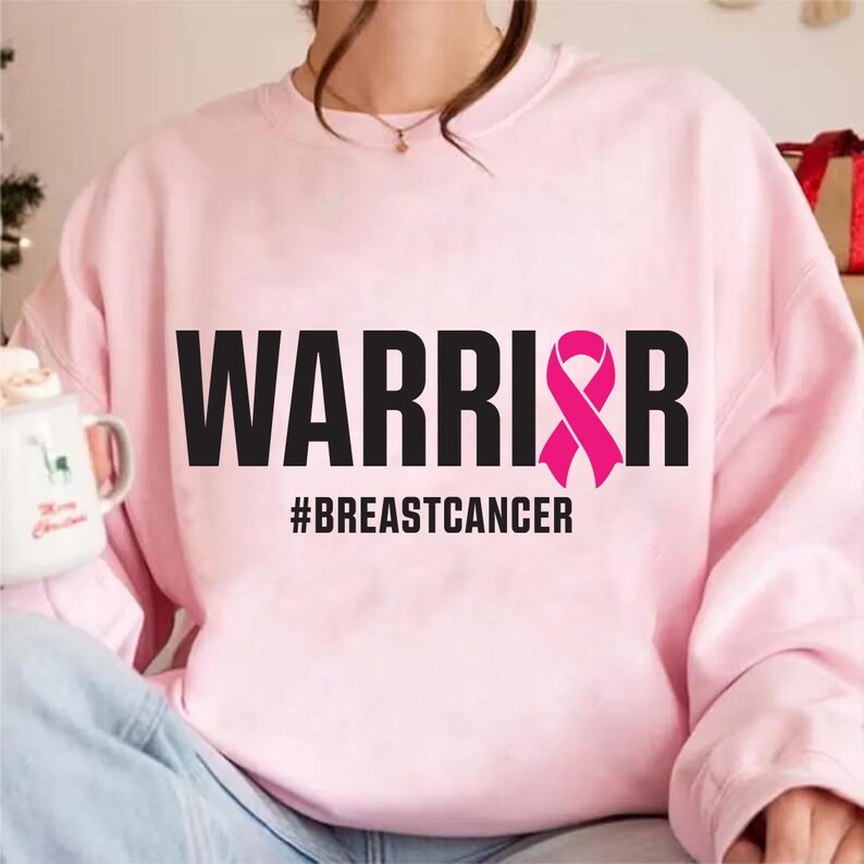 May include: A light pink sweatshirt with the word "WARRIOR" in black block letters and a pink ribbon graphic. Below the word is the hashtag "#BREASTCANCER". The sweatshirt is a crew neck style.