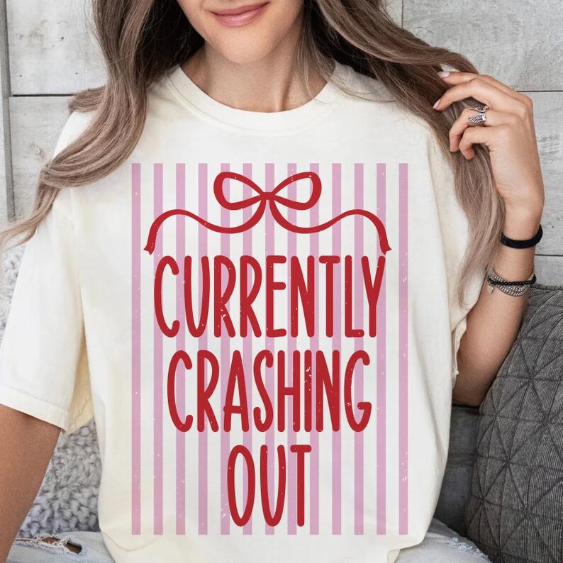 May include: Cream-colored t-shirt with a red bow and the words "CURRENTLY CRASHING OUT" in a bold, red font. The text is set against a background of vertical pink and white stripes. The shirt has a relaxed fit.