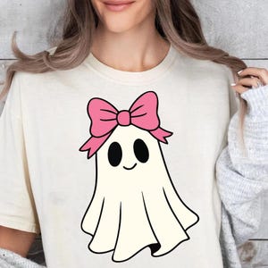 K&ouml;nnte beinhalten: Cremefarbenes T-Shirt mit einem Cartoon-Geist mit rosa Schleife. Der Geist hat schwarze ovale Augen und einen l&auml;chelnden Mund. Das Design ist mittig platziert und bietet eine verspielte und skurrile &Auml;sthetik.