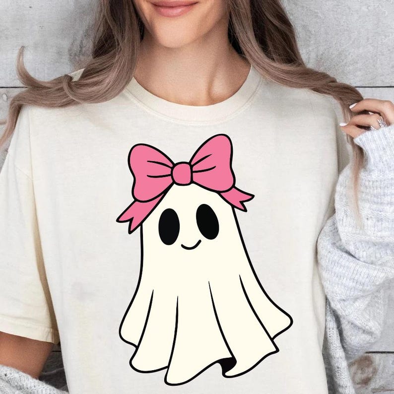 K&ouml;nnte beinhalten: Cremefarbenes T-Shirt mit einem Cartoon-Geist mit rosa Schleife. Der Geist hat schwarze ovale Augen und einen l&auml;chelnden Mund. Das Design ist mittig platziert und bietet eine verspielte und skurrile &Auml;sthetik.