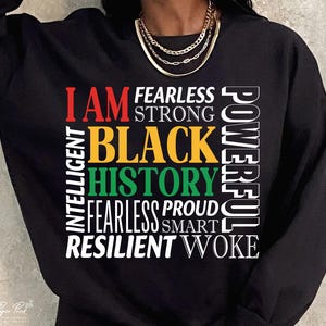 Peut inclure: Sweat-shirt noir avec les mots "I AM BLACK HISTORY" en rouge, jaune, vert et blanc. D'autres mots incluent "Fearless", "Strong", "Intelligent", "Proud", "Resilient" et "Woke". Un collier doré est visible.