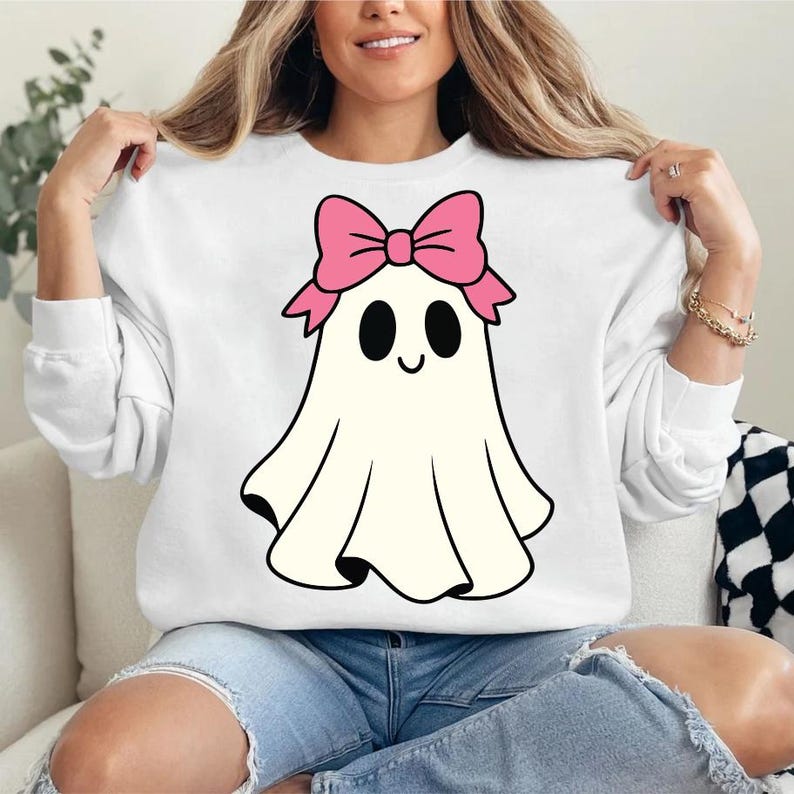 K&ouml;nnte beinhalten: Wei&szlig;es Sweatshirt mit einem Cartoon-Geist-Design. Der Geist hat eine rosa Schleife, schwarze Augen und einen l&auml;chelnden Mund. Das Design befindet sich auf der Vorderseite des lang&auml;rmeligen Oberteils. Das Model tr&auml;gt eine blaue Jeans.