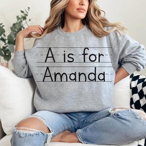 May include: A gray sweatshirt with the text "A is for Amanda" in black. The text is written in a handwritten style, with lines underneath. The sweatshirt is a crew neck style.