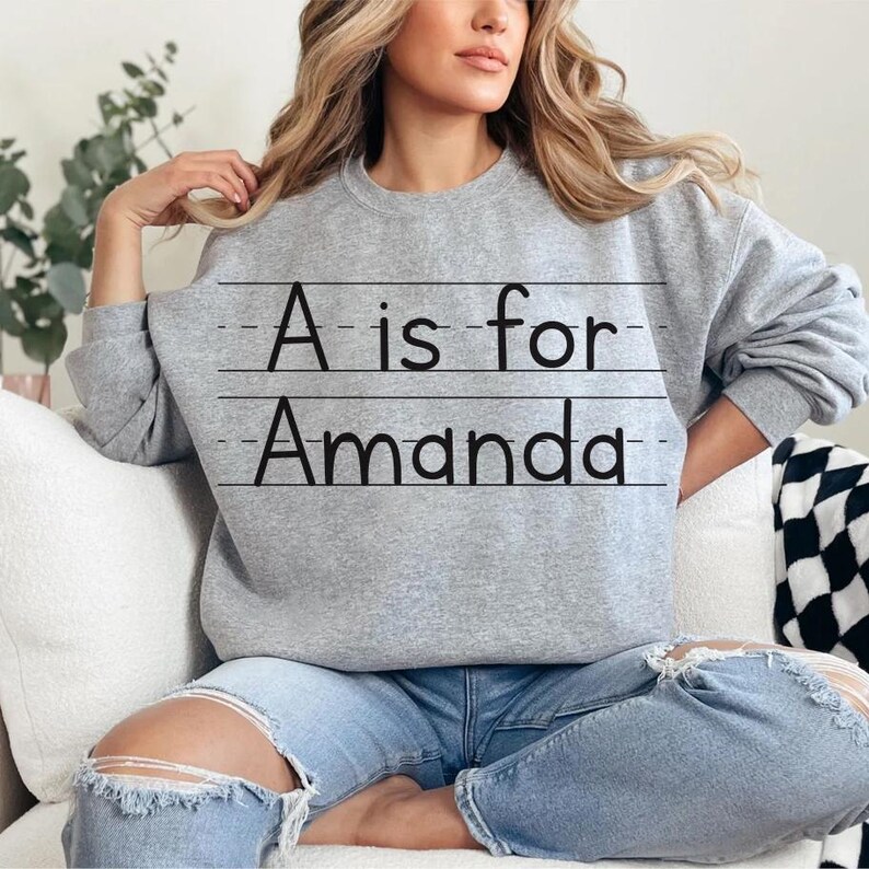 May include: A gray sweatshirt with the text "A is for Amanda" in black. The text is written in a handwritten style, with lines underneath. The sweatshirt is a crew neck style.