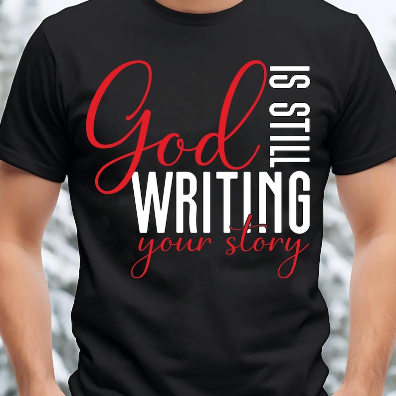 May include: Black t-shirt with the text "God is still writing your story" in red and white. The words "God" and "story" are in a cursive font. The shirt is a crew neck.