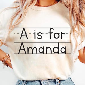 May include: Cream-colored t-shirt with the text "A is for Amanda" in black, written on lined paper. The shirt is a casual style, with a round neckline and short sleeves.