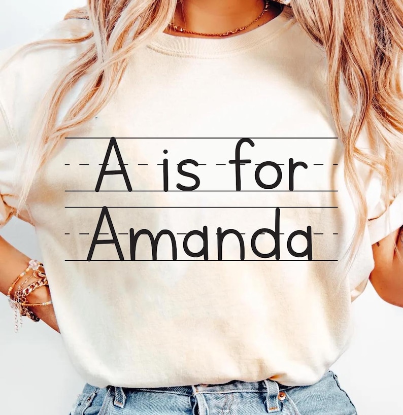 May include: Cream-colored t-shirt with the text "A is for Amanda" in black, written on lined paper. The shirt is a casual style, with a round neckline and short sleeves.