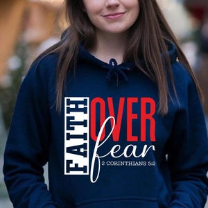 May include: Navy blue hooded sweatshirt with the words "FAITH OVER FEAR" in white and red lettering. The text "2 Corinthians 5:2" is below the word "fear". The sweatshirt has a drawstring and a front pocket.