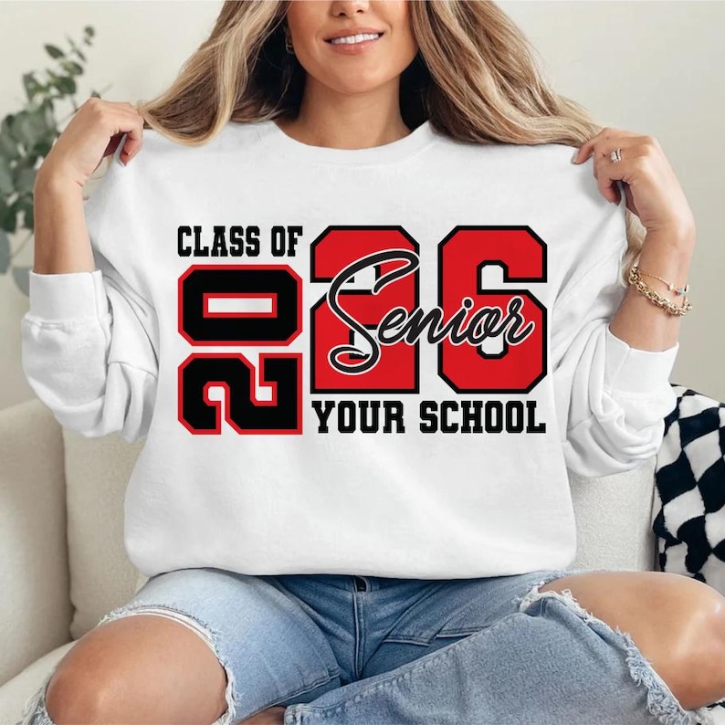 Pode incluir: Camisola branca com o texto "CLASS OF 26 SENIOR YOUR SCHOOL" em letras pretas e vermelhas. A camisola tem mangas compridas e gola redonda. O design est&aacute; centrado na frente.