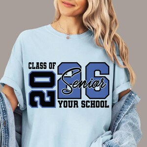 Pode incluir: Camiseta azul clara com o texto "CLASS OF 26 Senior YOUR SCHOOL" em preto e azul. A camiseta &eacute; de gola redonda e est&aacute; sendo usada por uma pessoa.