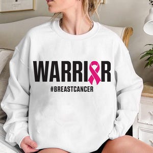 May include: White sweatshirt with the word "WARRIOR" in bold black letters and a pink ribbon graphic. The hashtag "#BREASTCANCER" is printed below the word "WARRIOR". The sweatshirt is a crew neck style.