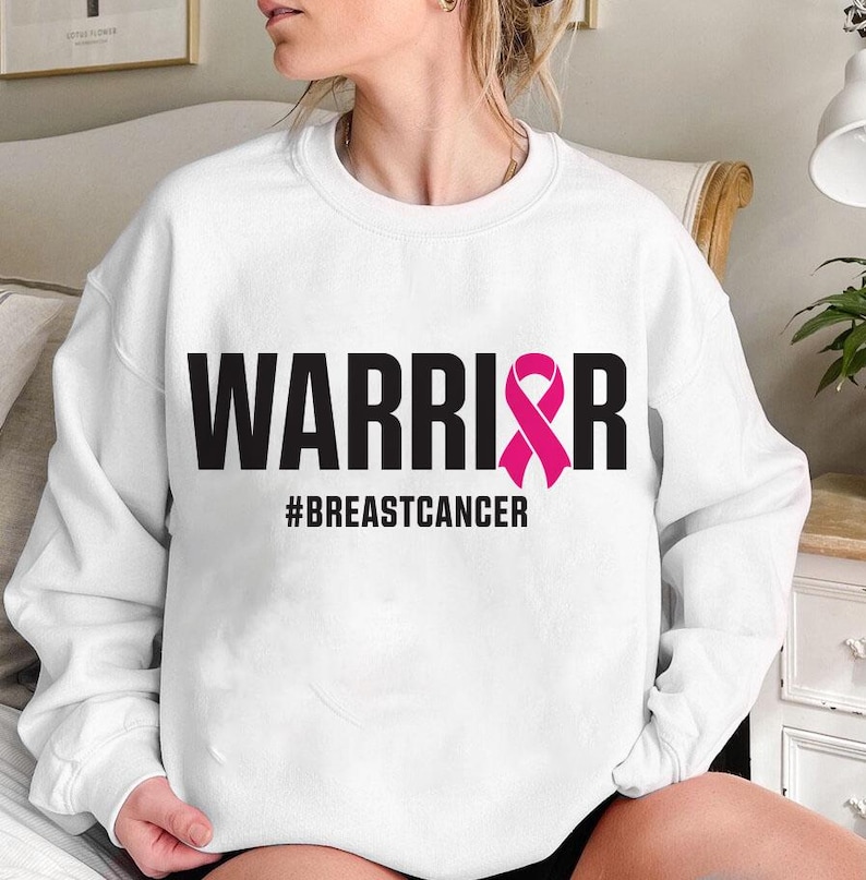May include: White sweatshirt with the word "WARRIOR" in bold black letters and a pink ribbon graphic. The hashtag "#BREASTCANCER" is printed below the word "WARRIOR". The sweatshirt is a crew neck style.