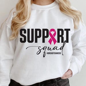 May include: White sweatshirt with the words "SUPPORT SQUAD" in black block letters and a pink ribbon graphic. The word "squad" is in a cursive font, with the hashtag "#BREASTCANCER" below.