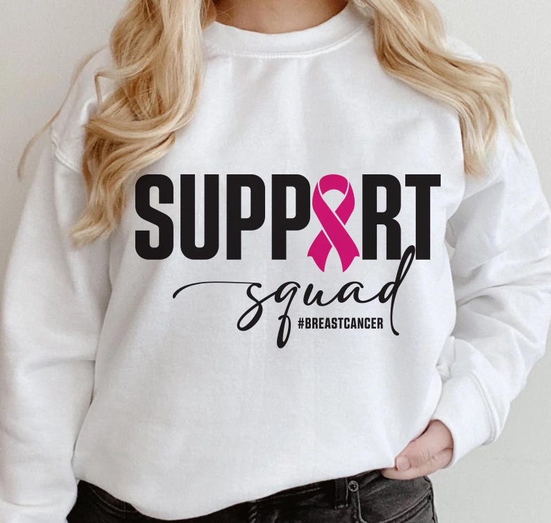May include: White sweatshirt with the words "SUPPORT SQUAD" in black block letters and a pink ribbon graphic. The word "squad" is in a cursive font, with the hashtag "#BREASTCANCER" below.